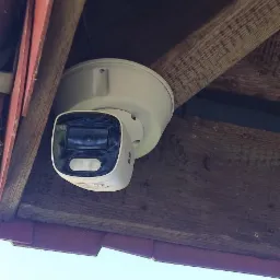 Biała kamera monitoringu zamontowana pod drewnianym zadaszeniem z czerwoną krawędzią, widok z dołu na błękitne niebo.