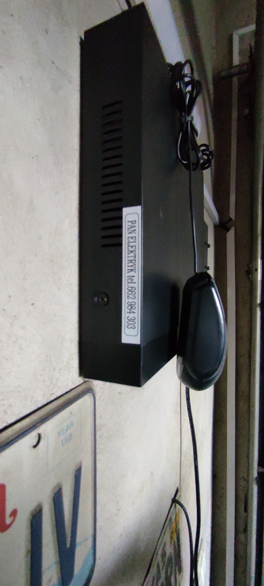 Czarna centrala alarmowa z naklejoną etykietą firmy 'PAN ELEKTRYK' i numerem telefonu, zamontowana na jasnej ścianie obok tablic rejestracyjnych.