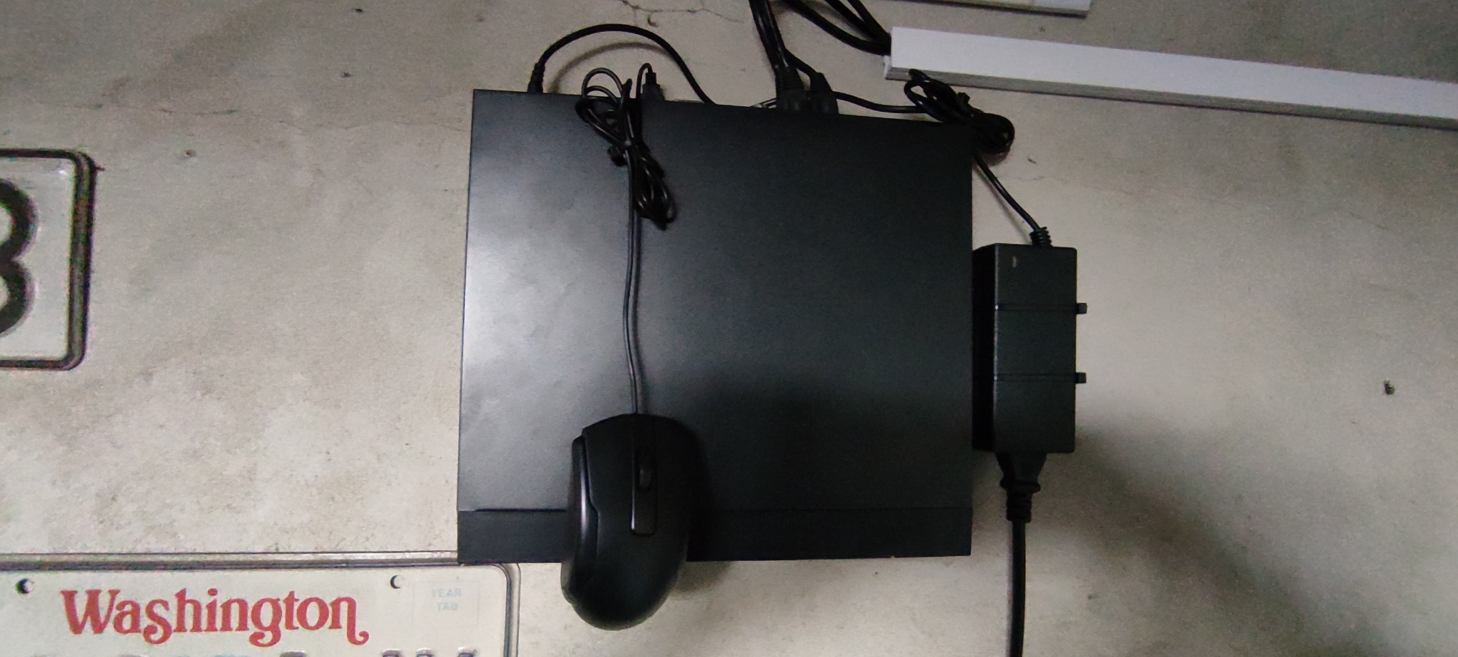 Czarna centrala alarmowa z podłączonym zasilaczem i myszką komputerową zawieszona na szarej ścianie, widoczny fragment tablicy rejestracyjnej.