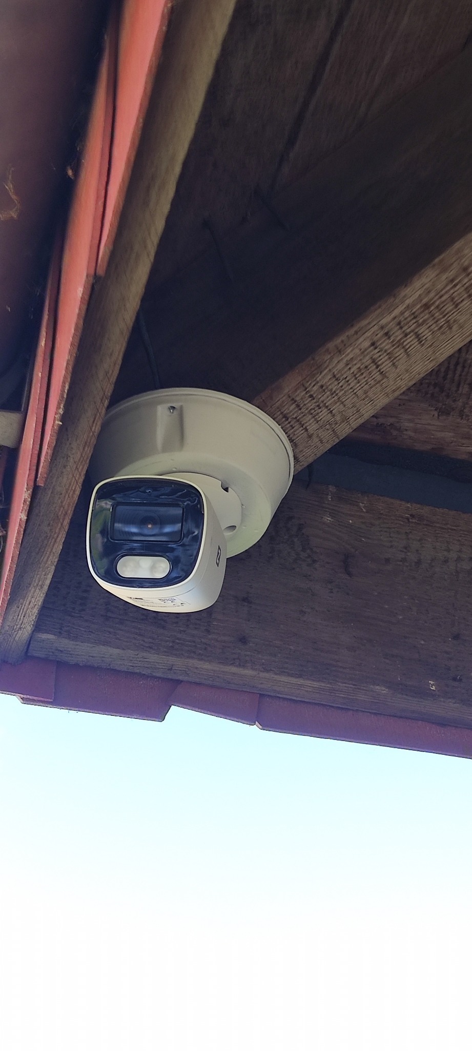 Biała kamera monitoringu zamontowana pod drewnianym zadaszeniem z czerwoną krawędzią, widok z dołu na błękitne niebo.