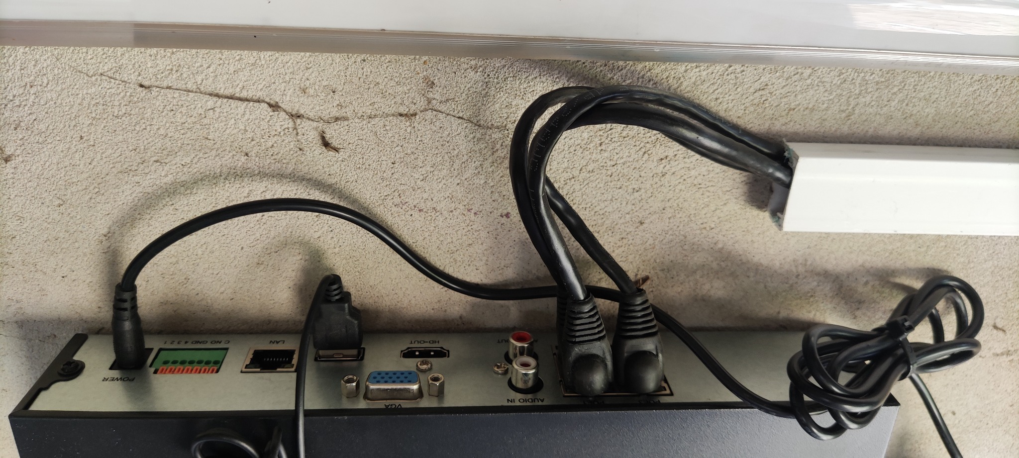 Widok z tyłu rejestratora monitoringu z podłączonymi kablami: zasilanie, LAN, HDMI, VGA oraz audio, zamocowanego na szorstkiej ścianie w pobliżu białej listwy przypodłogowej.