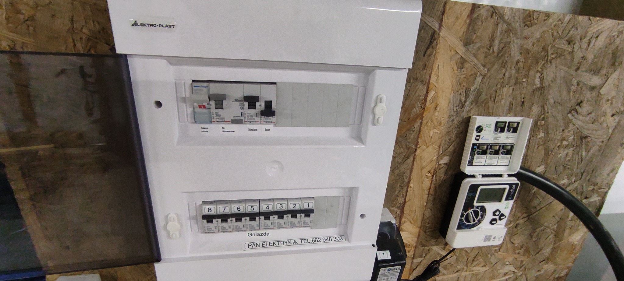 Biała skrzynka rozdzielcza Elektro-Plast z widocznymi bezpiecznikami i listwą zaciskową, zamontowana na ścianie z płyty OSB, obok automatyczny sterownik nawadniania c-dial.