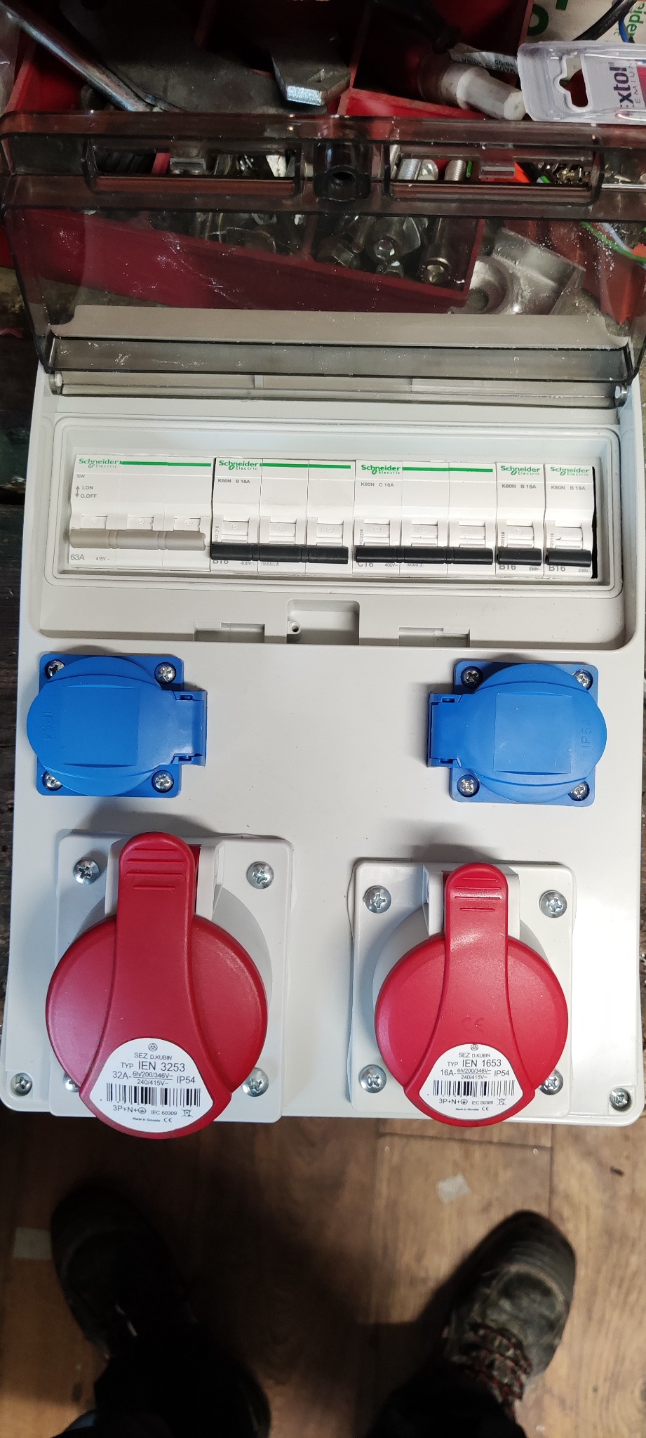 Rozdzielnica elektryczna z wyłącznikami różnicowoprądowymi Schneider Electric i gniazdami siłowymi (czerwone i niebieskie), widoczne etykiety gniazd z oznaczeniami IEN 3253 i IEN 1653, częściowo...