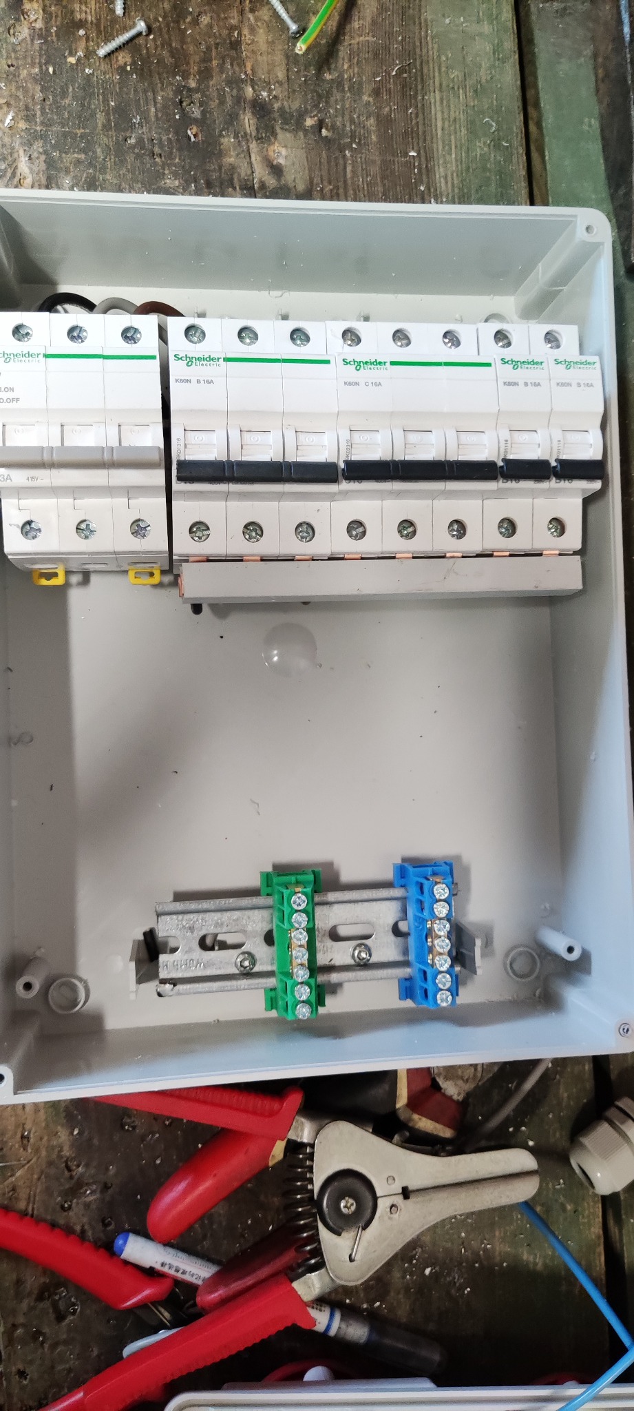 Otwarta skrzynka elektryczna z zamontowanymi bezpiecznikami Schneider Electric K60N, listwami zaciskowymi w kolorze zielonym i niebieskim oraz narzędziami elektrycznymi w tle.