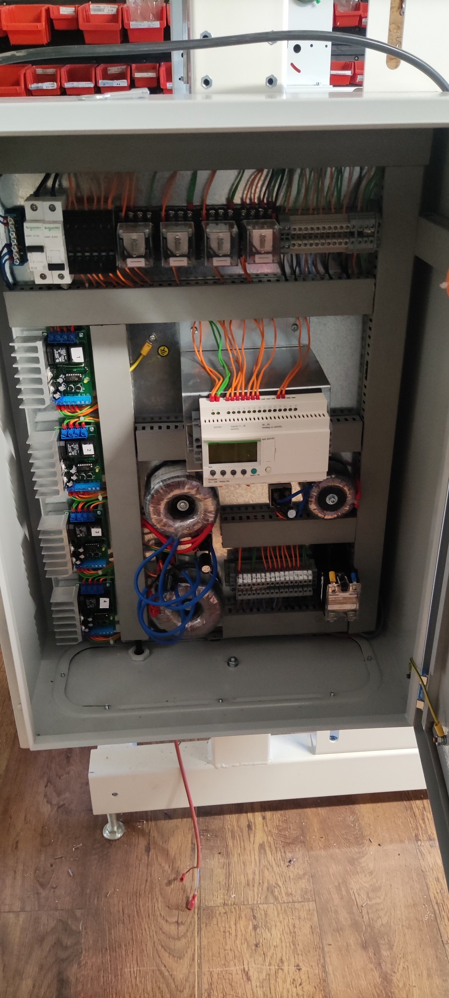 Otwarta szafa sterownicza z widocznymi transformatorami toroidalnymi, sterownikiem PLC, bezpiecznikami i okablowaniem w kolorach pomarańczowym, zielonym i niebieskim.