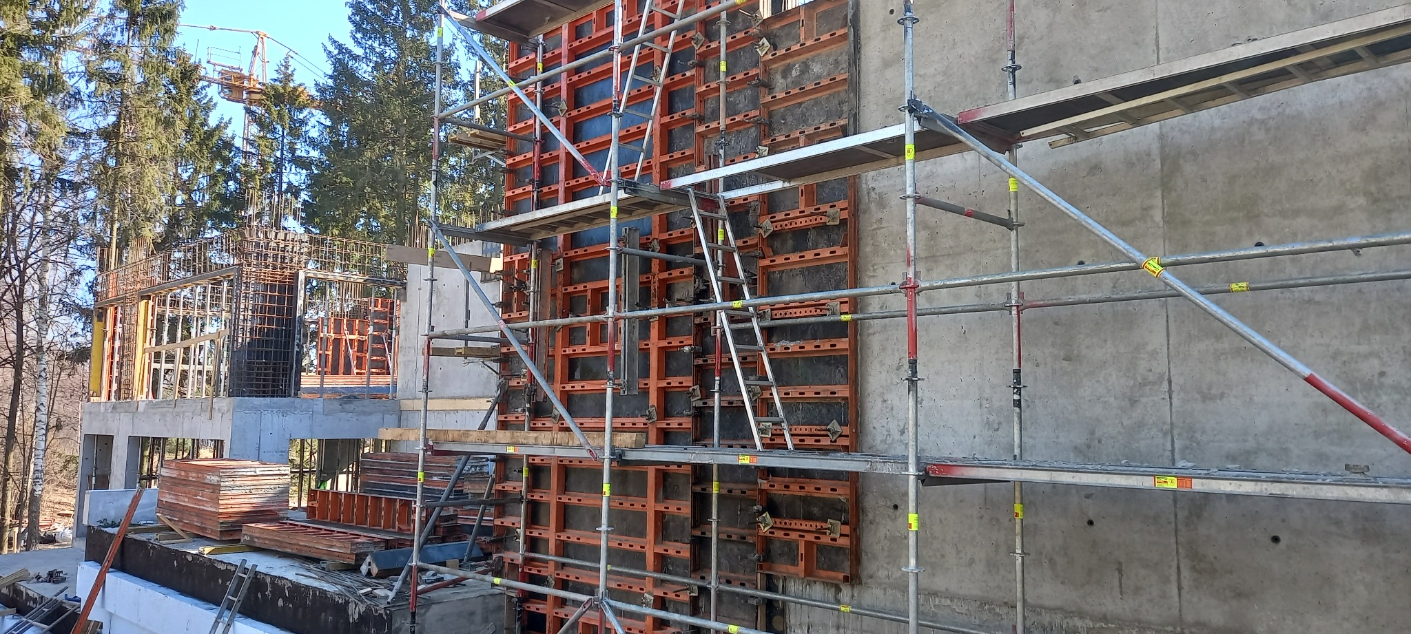 Szare rusztowania przy betonowej ścianie z zamontowanym pomarańczowym szalunkiem modułowym na budowie, w tle drzewa i żuraw budowlany.