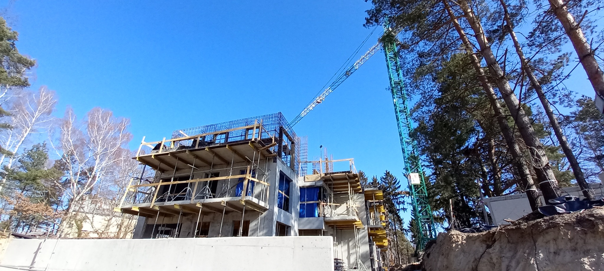 Budowa domu wielorodzinnego w stanie surowym otwartym, widoczny żuraw budowlany na tle błękitnego nieba i drzew