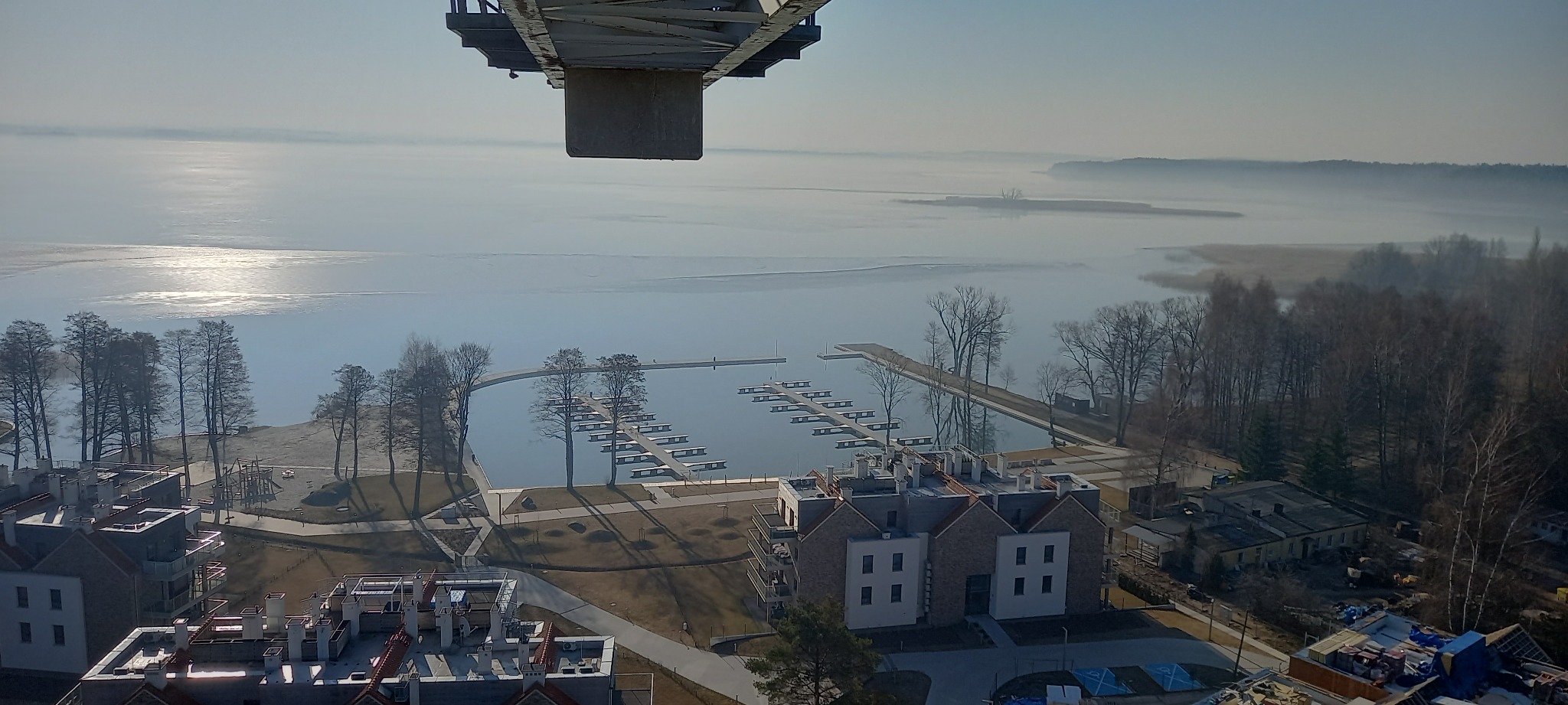 Panorama z dźwigu budowlanego: widok na jezioro z pomostami, częściowo ukończone budynki mieszkalne z czerwonymi dachami oraz plac zabaw.