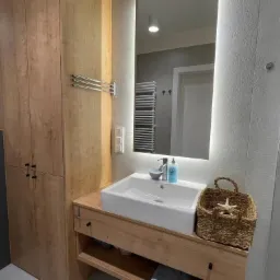 Nowoczesna łazienka z drewnianą szafką pod umywalką, podświetlanym lustrem i dekoracyjnym koszem z gwiazdą morską, widoczny fragment szafy z drewnianymi frontami.