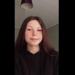 Młoda kobieta z długimi brązowymi włosami w czarnej bluzie z kapturem, uśmiechnięta, widoczna od ramion w górę, w tle sufit z lampą.