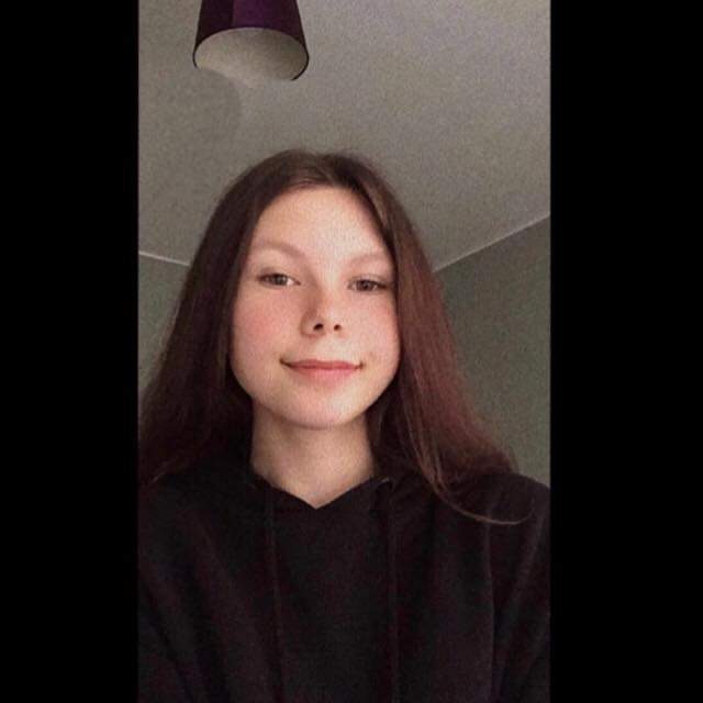 Młoda kobieta z długimi brązowymi włosami w czarnej bluzie z kapturem, uśmiechnięta, widoczna od ramion w górę, w tle sufit z lampą.