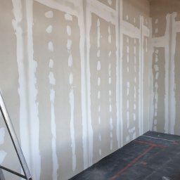 Krispol-Bud - Ściana w trakcie przygotowania do malowania, z widocznymi pasami świeżo nałożonej gładzi szpachlowej na łączeniach płyt kartonowo-gipsowych, częściowo zakryta rusztowaniem z czarną matą...