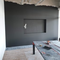 Krispol-Bud - Częściowo pomalowana na ciemny kolor ściana biura z wnęką na telewizor, widoczne białe rury pod sufitem, zabezpieczona folią roboczą powierzchnia stołu z narzędziami i niedopitą kawą, drewniana...