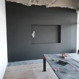 Częściowo pomalowana na ciemny kolor ściana biura z wnęką na telewizor, widoczne białe rury pod sufitem, zabezpieczona folią roboczą powierzchnia stołu z narzędziami i niedopitą kawą, drewniana...