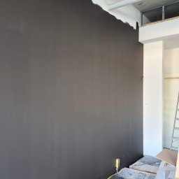 Krispol-Bud - Świeżo pomalowana na szaro ściana w pomieszczeniu w trakcie wykańczania, widoczne zabezpieczenia podłogi i niedomalowane fragmenty przy suficie.