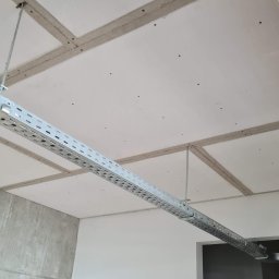 Krispol-Bud - Sufit z płyt gipsowo-kartonowych ze szpachlowanymi łączeniami, widoczna metalowa korytka kablowa podwieszona do sufitu, surowa ściana betonowa w rogu pomieszczenia.