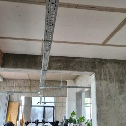 Krispol-Bud - Surowy beton i sufit z płyt gipsowo-kartonowych z zamontowanymi metalowymi korytkami kablowymi w przestrzeni biurowej w trakcie prac wykończeniowych.