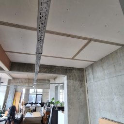 Krispol-Bud - Montaż instalacji pod sufitem podwieszanym w surowym wnętrzu biurowym, widoczne metalowe korytka kablowe, betonowe ściany i drewniana podłoga, w tle pracownik i biurka.