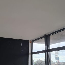 Krispol-Bud - Wnętrze po świeżym malowaniu na biało, widoczna czarna ściana, sufit i okno z widokiem na port z dźwigiem.