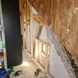 Krispol-Bud - Wnętrze w trakcie prac remontowych: drewniana konstrukcja szkieletowa ścianki działowej, odsłonięte fragmenty starego muru i instalacji elektrycznej, narzędzia i materiały budowlane na podłodze.