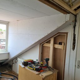 Krispol-Bud - Wnętrze w trakcie remontu: ściany pokryte płytami gipsowo-kartonowymi, widoczne narzędzia i materiały budowlane na komodzie, odsłonięta instalacja elektryczna w ścianie.