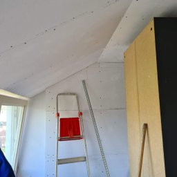 Krispol-Bud - Poddasze w trakcie zabudowy płytami gipsowo-kartonowymi, widoczna drabina aluminiowa z czerwonymi elementami, profil metalowy, fragment szafy i okno dachowe.