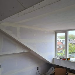Krispol-Bud - Poddasze w trakcie prac wykończeniowych: ściany pokryte szpachlą, widoczne łączenia płyt gipsowo-kartonowych, poziomica, wiadro z masą szpachlową i narzędzia na podłodze, okno z widokiem...