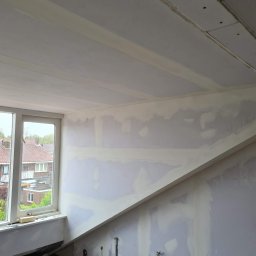 Krispol-Bud - Wnętrze poddasza w trakcie szpachlowania z widocznymi łączeniami płyt gipsowo-kartonowych, z oknem wychodzącym na zabudowę jednorodzinną.