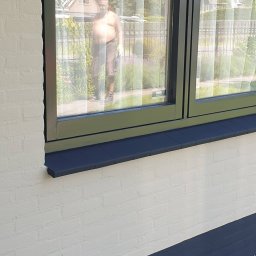 Krispol-Bud - Biała ściana z cegły z pomalowaną na granatowo podmurówką i parapetem, widok przez okno na mężczyznę w ogrodzie.