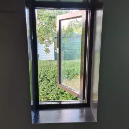 Świeżo pomalowane okno z widokiem na zieleń ogrodu, ciemnoszara rama okienna kontrastuje z kolorem ściany i otwartym skrzydłem okna.