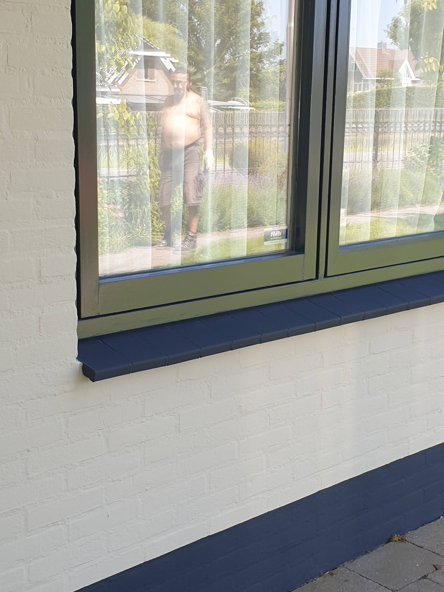 Biała ściana z cegły z pomalowaną na granatowo podmurówką i parapetem, widok przez okno na mężczyznę w ogrodzie.