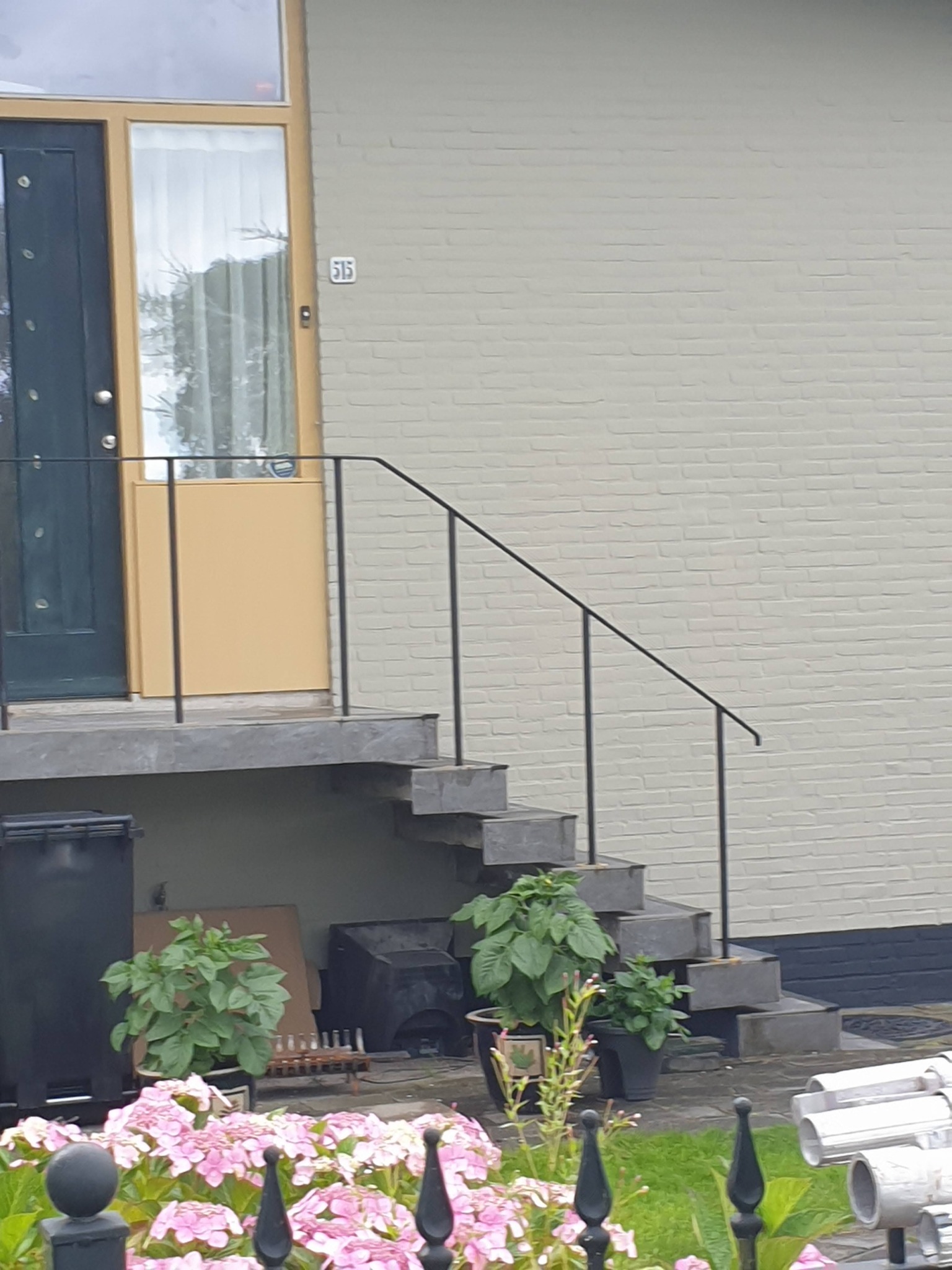 Wejście do domu z betonowymi schodami i czarną balustradą, jasna elewacja z numerem 515, donice z zielenią i różowe hortensje w pierwszym planie.