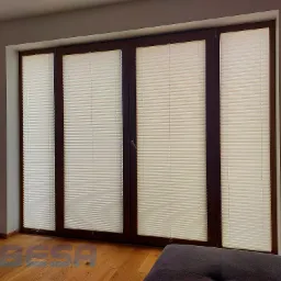 Czteroskrzydłowe okno z brązowymi ramami wyposażone w beżowe plisy, widoczne z wnętrza pomieszczenia z parkietem i robotem sprzątającym.