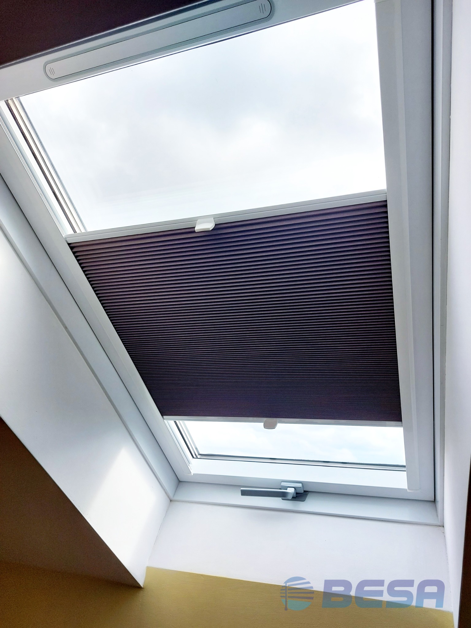 Częściowo zasłonięte okno dachowe z plisowaną, ciemnofioletową roletą, widok z dołu, jasne niebo w tle, fragment ściany z logo firmy.