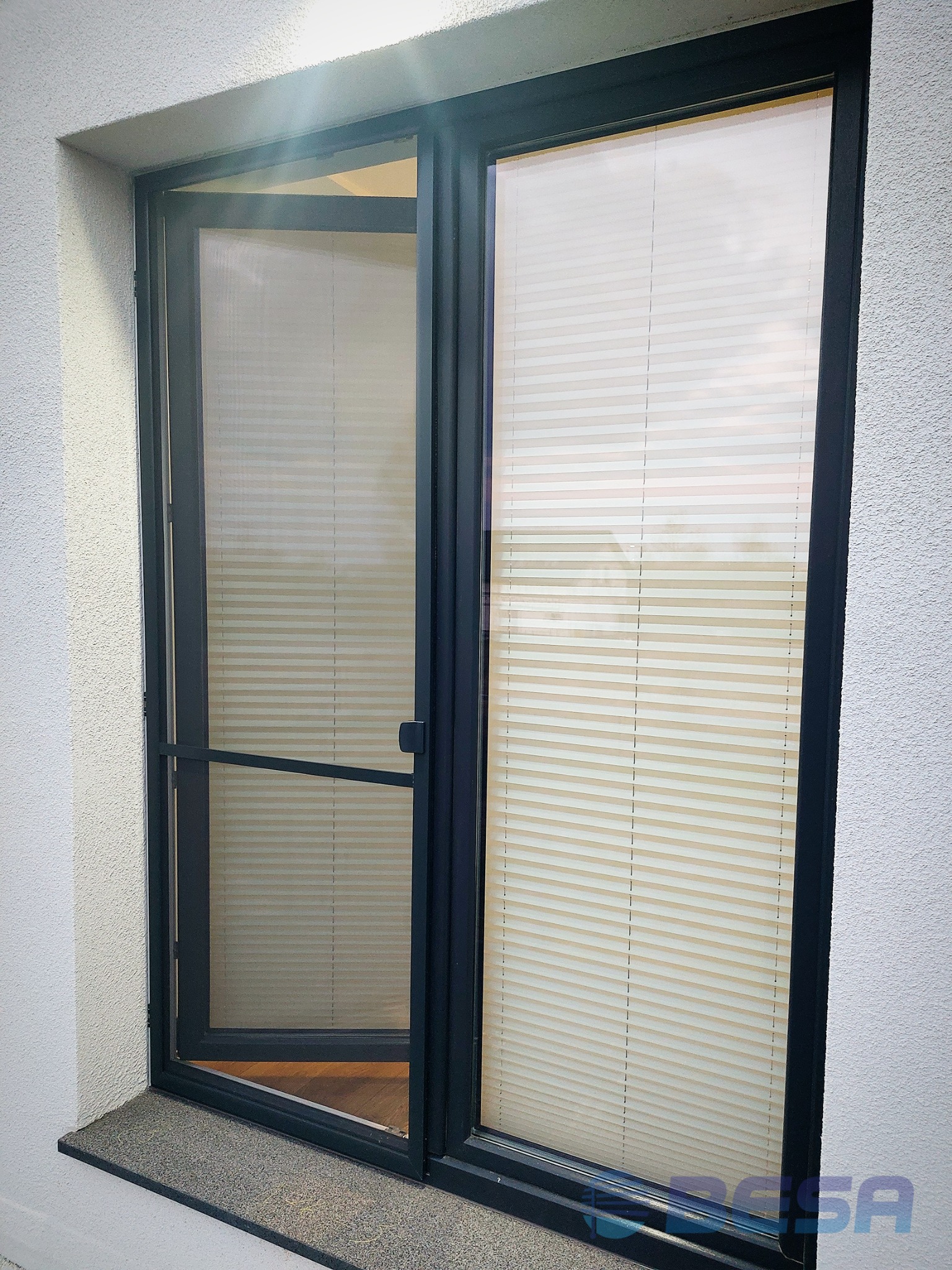 Otwierane okno z czarną ramą i plisami w kolorze ecru, widok z zewnątrz na elewację o szorstkiej fakturze.