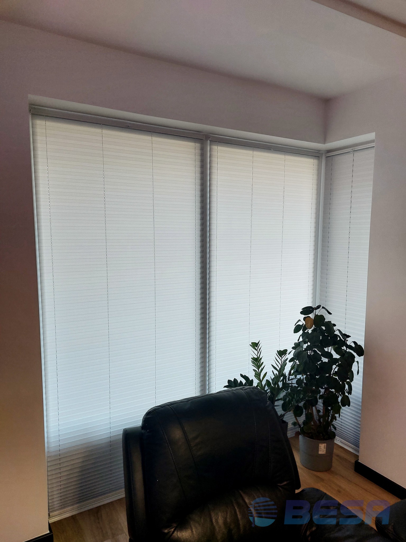 Jasne, aluminiowe żaluzje poziome zamontowane w oknie w Chojnicach, widoczne zza skórzanego fotela i doniczkowych roślin, tworzące stonowane tło.