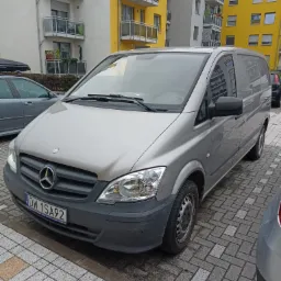 Srebrny Mercedes-Benz Vito zaparkowany na brukowanej powierzchni przed budynkiem mieszkalnym z żółtymi i białymi balkonami.