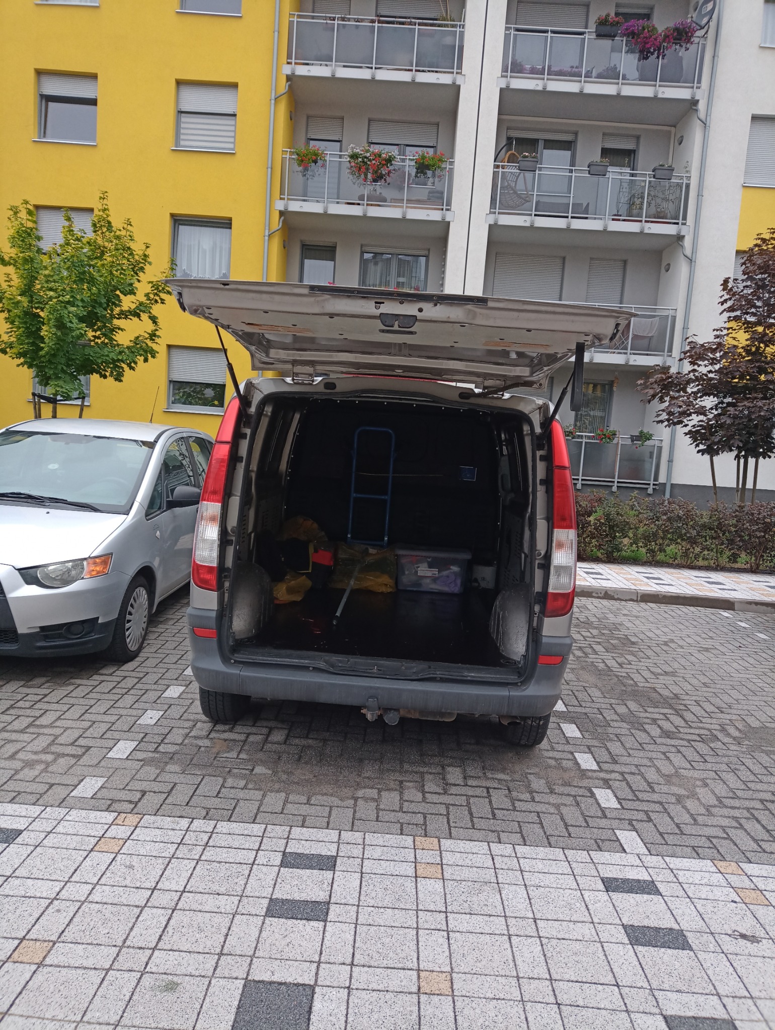 Tył srebrnego vana z otwartą klapą bagażnika, częściowo załadowanego narzędziami i skrzynką, zaparkowanego na kostce brukowej przed budynkiem mieszkalnym.
