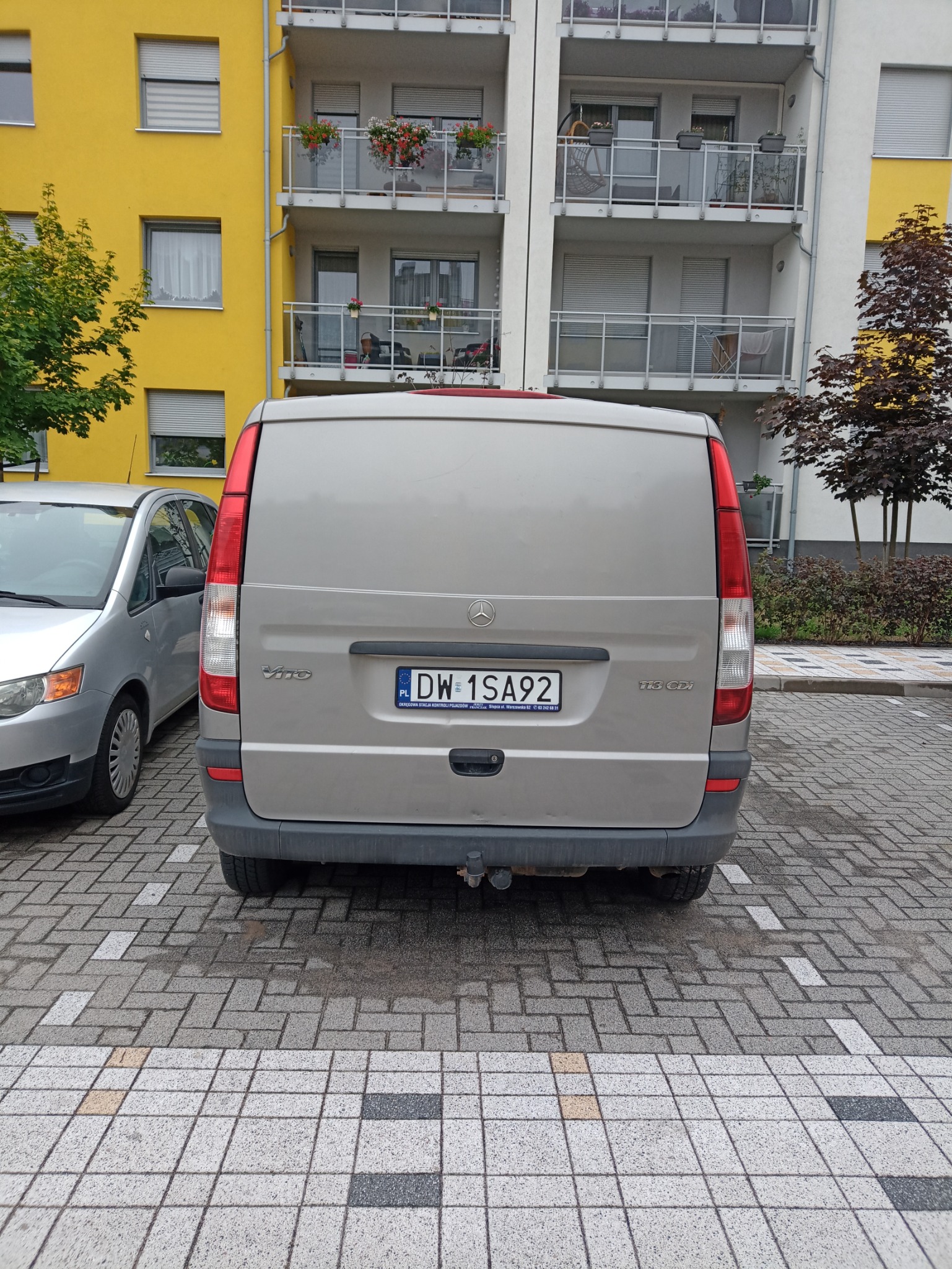 Tył szarego busa Mercedes Vito zaparkowanego na kostce brukowej, widoczny numer rejestracyjny DW 1SA92, w tle budynek mieszkalny z balkonami.