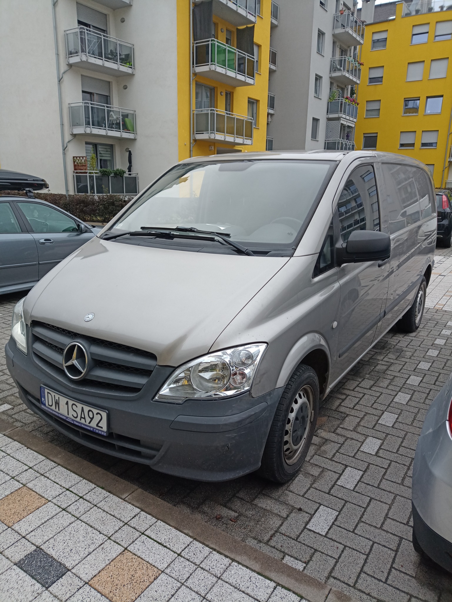 Srebrny Mercedes-Benz Vito zaparkowany na brukowanej powierzchni przed budynkiem mieszkalnym z żółtymi i białymi balkonami.