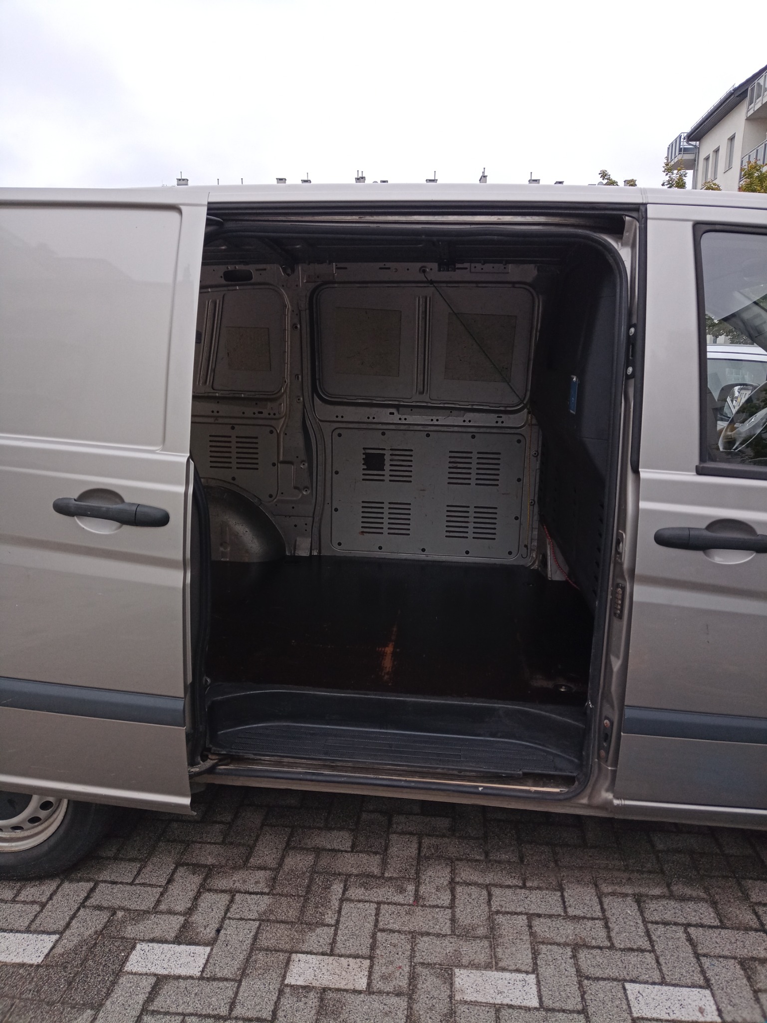 Wnętrze pustego busa transportowego z otwartymi drzwiami bocznymi, widok na podłogę i metalowe panele boczne z otworami wentylacyjnymi.