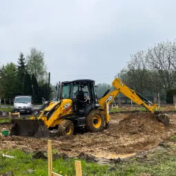 Żółta koparko-ładowarka JCB podczas prac ziemnych na budowie, widoczne drewniane szalunki fundamentów, w tle biały bus i drzewa.