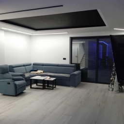 W.A.G Team - Nowoczesny salon z podwieszanym dwupoziomowym sufitem z oświetleniem LED, jasnymi panelami podłogowymi i narożną sofą w odcieniach niebieskiego.