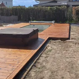Świeżo polakierowany taras z drewna otaczający basen i jacuzzi, widoczne ślady lakieru i niedokończone prace ziemne w ogrodzie.