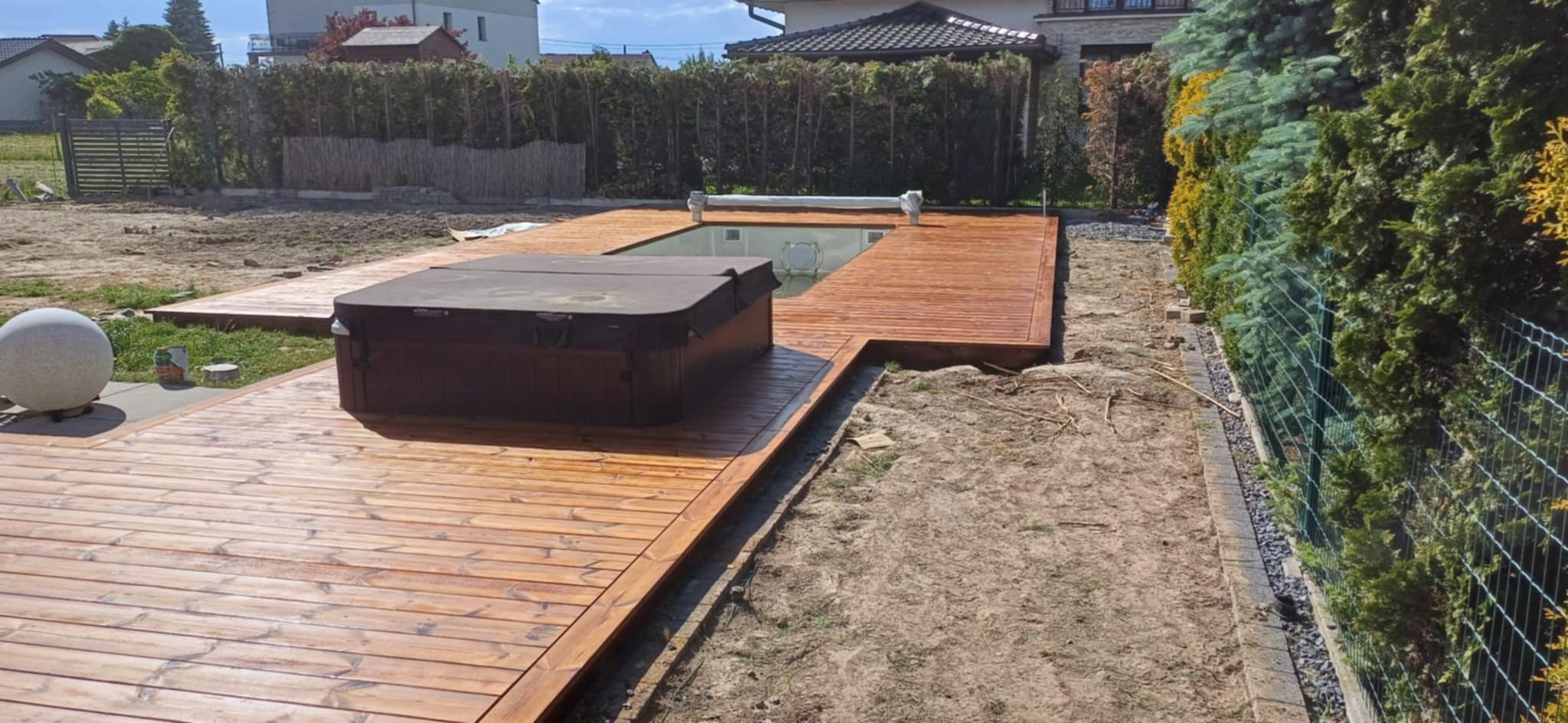 Świeżo polakierowany taras z drewna otaczający basen i jacuzzi, widoczne ślady lakieru i niedokończone prace ziemne w ogrodzie.