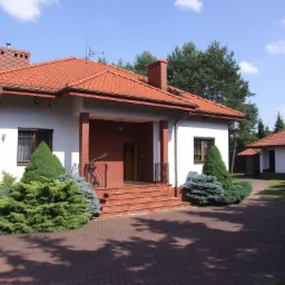 Dom jednorodzinny z czerwoną dachówką, ceglanym wejściem i ozdobnymi krzewami iglastymi na podjeździe z kostki brukowej.