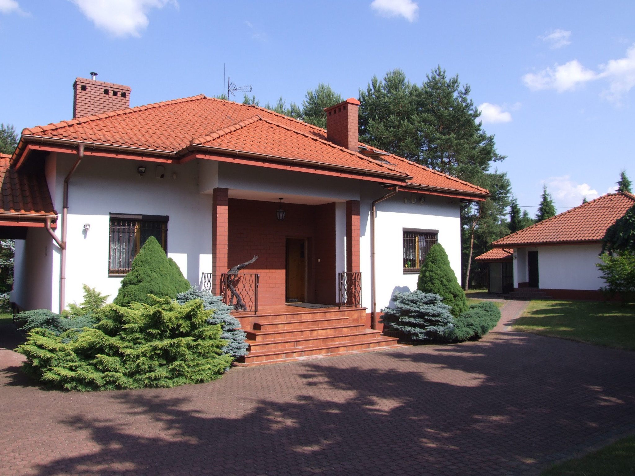 Dom jednorodzinny z czerwoną dachówką, ceglanym wejściem i ozdobnymi krzewami iglastymi na podjeździe z kostki brukowej.