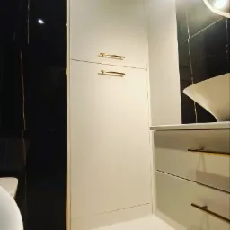 Nowoczesna łazienka z szafką w zabudowie, złotymi uchwytami i czarną, marmurkową ścianą w tle.