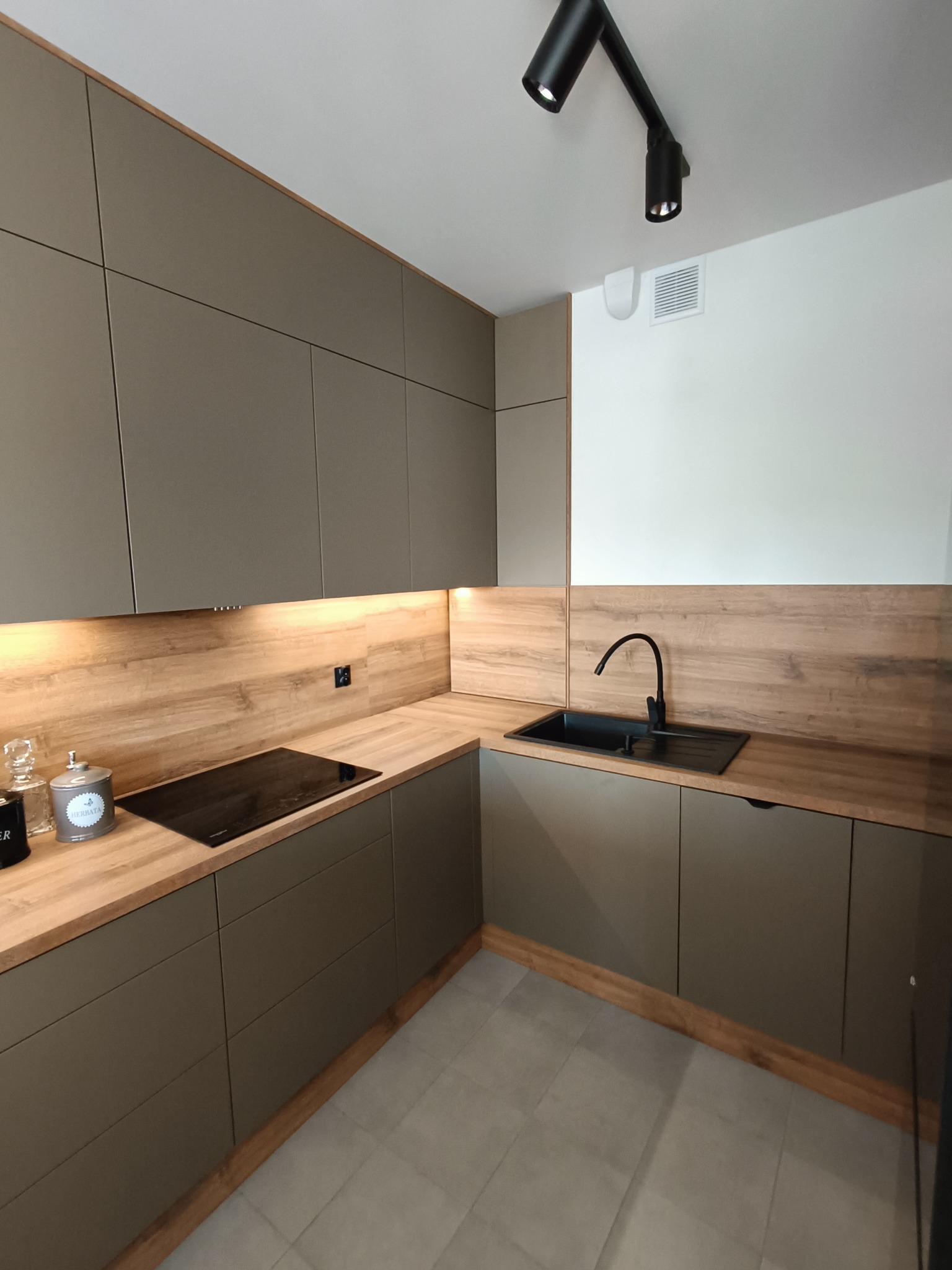 Nowoczesna kuchnia z szarymi frontami i drewnianym blatem w Zabrzu. Minimalistyczny design, czarny zlew i płyta indukcyjna. Oświetlenie punktowe dodaje elegancji.