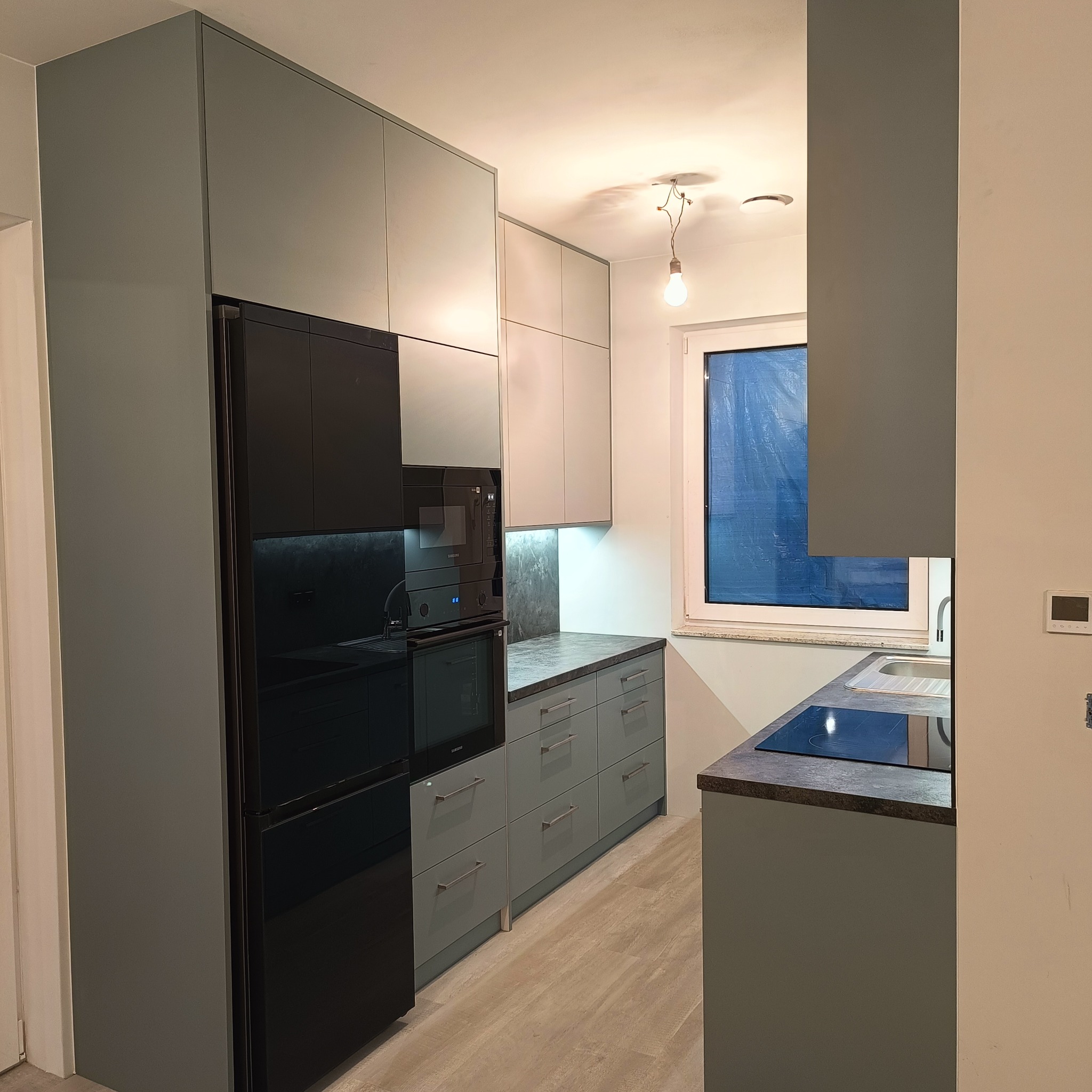 Nowoczesna kuchnia w odcieniach szarości z czarną lodówką i piekarnikiem. Minimalistyczne szafki, drewnopodobna podłoga i okno z widokiem na zewnątrz.